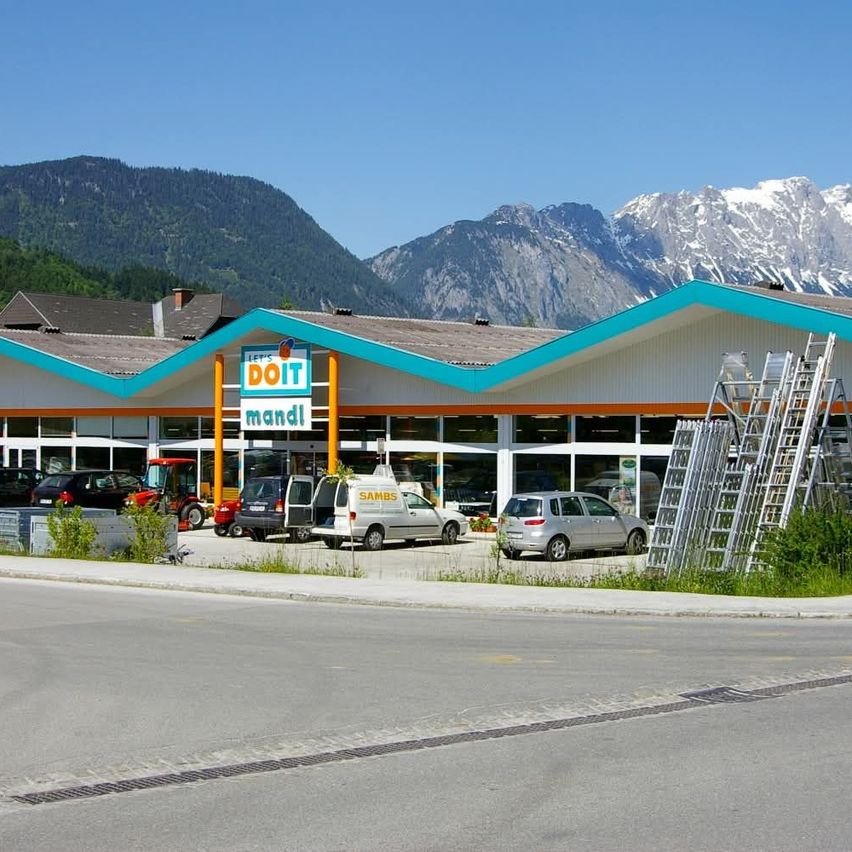 Ein Baumarkt steht vor schneebedeckten Bergen. Mehrere Autos und ein Lieferwagen sind draußen geparkt. Eine Leiter ist gegen das Gebäude gelehnt.