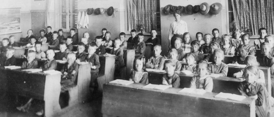 Ein Schwarz-Weiß-Foto einer Klasse mit Kindern an ihren Schreibtischen. Der Lehrer steht vorne.
