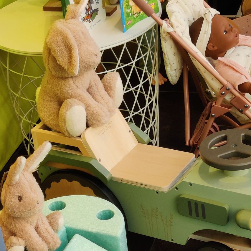 Zwei Plüschhasen, einer sitzt auf einem Spielzeugauto und der andere auf einem kleinen Block. Im Hintergrund eine Puppe in einem Kinderwagen und ein kleiner Tisch mit einem Buch.