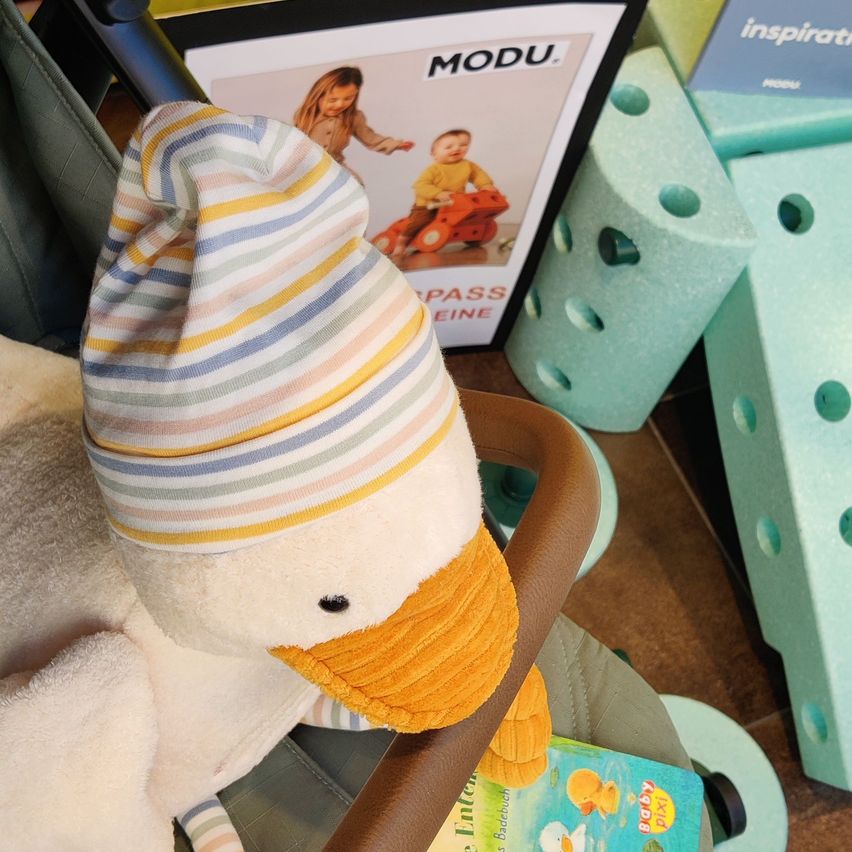 Nahaufnahme eines weißen Enten-Plüschtiers, das auf dem Griff eines Kinderwagens sitzt, mit einer mehrfarbigen Mütze auf dem Kopf. Dahinter befinden sich verschiedene farbige Schaumstoffblöcke und ein Buch mit einem Bild eines Kindes, das ein Spielzeugauto fährt.