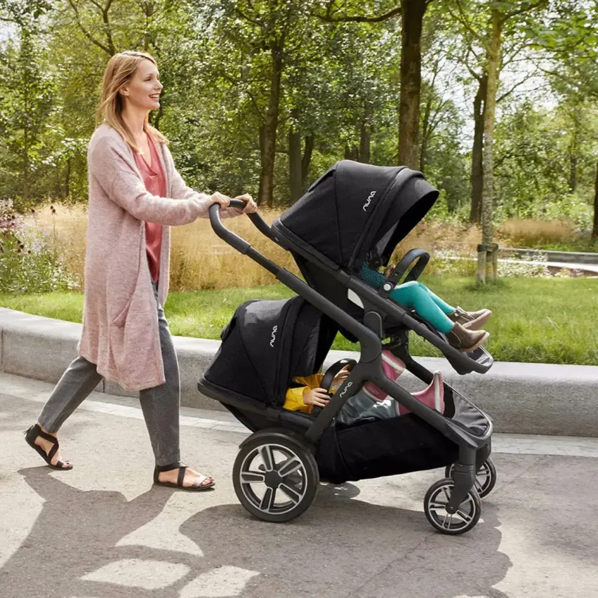 Eine Frau in einem rosa Cardigan schiebt einen schwarzen Kinderwagen mit zwei Kindern darin. Der Kinderwagen hat zwei Sitze, und die Kinder tragen Schuhe. Die Frau trägt Sandalen. Der Hintergrund ist eine grasbewachsene Fläche mit Bäumen.