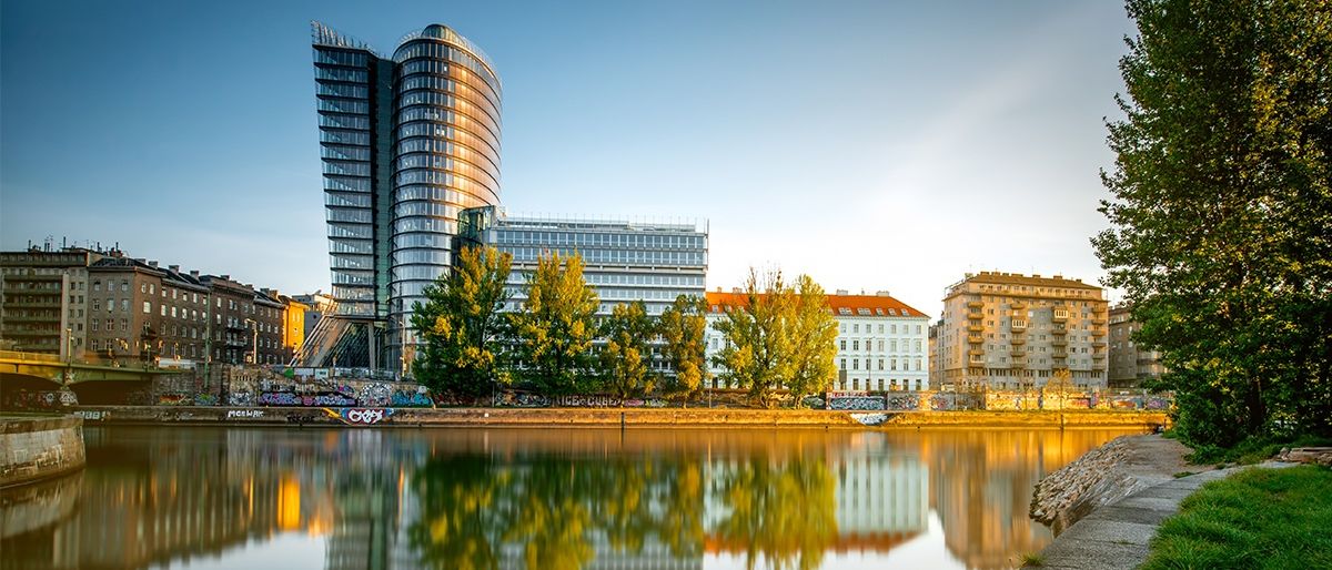 Ein modernes Gebäude mit Glasfassaden spiegelt sich auf der Wasseroberfläche wider, umgeben von Bäumen und anderen Gebäuden unter einem klaren Himmel.