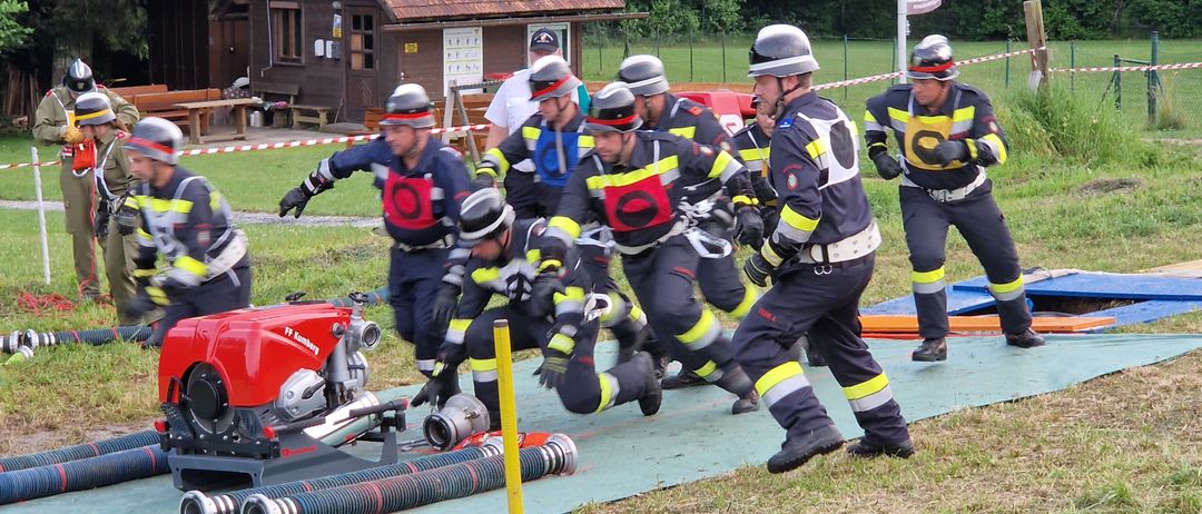 Eine Gruppe von Feuerwehrleuten in voller Ausrüstung nimmt an einer Trainingsübung auf einem Grasfeld teil. Sie rennen auf einen roten Feuerwehrwagen zu, an dem Schläuche und Ausrüstungsgegenstände ausgelegt sind.