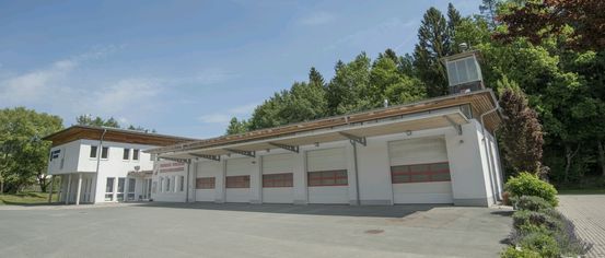 Ein weißes Gebäude mit einem braunen Dach hat vier Garagentore mit roten Einfassungen. Bäume sind im Hintergrund und der Himmel ist klar.