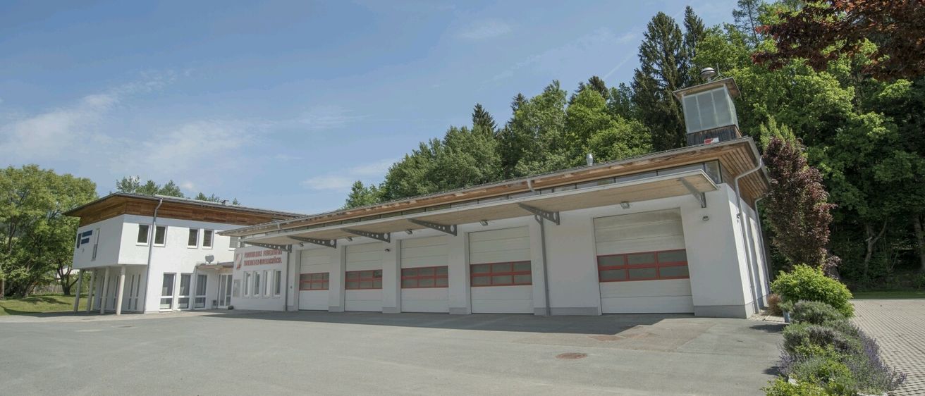 Ein weißes Gebäude mit einem braunen Dach hat vier Garagentore mit roten Einfassungen. Bäume sind im Hintergrund und der Himmel ist klar.