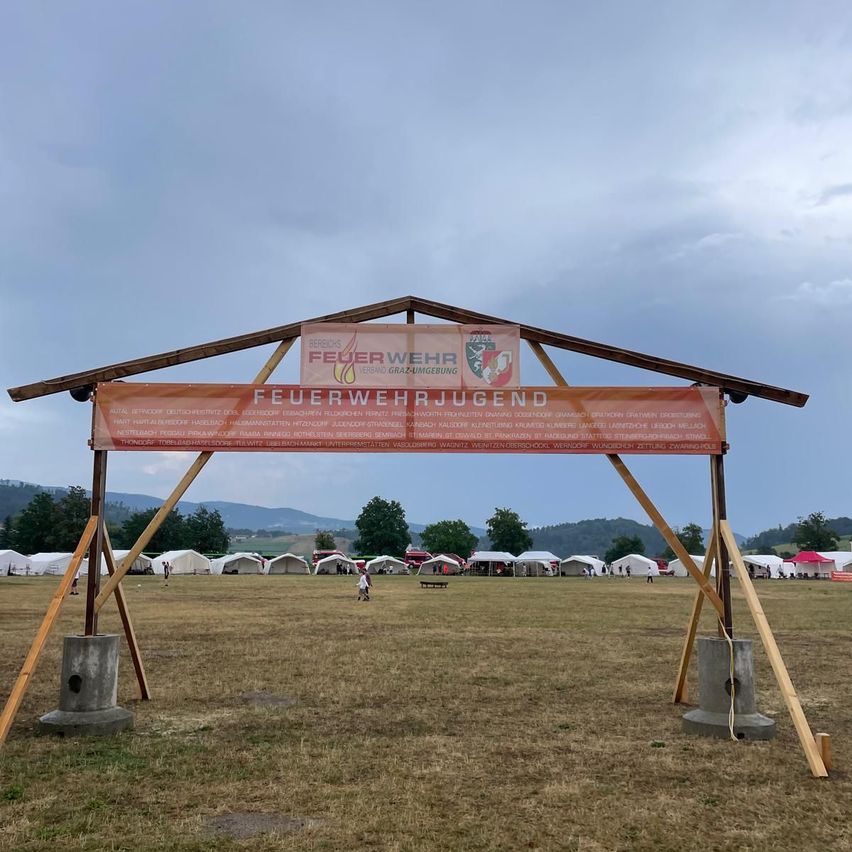 Ein hölzerner Bogen mit einem Banner, das 'Feuerwehrjugend' steht in einem offenen Feld mit Zelten und Menschen im Hintergrund.