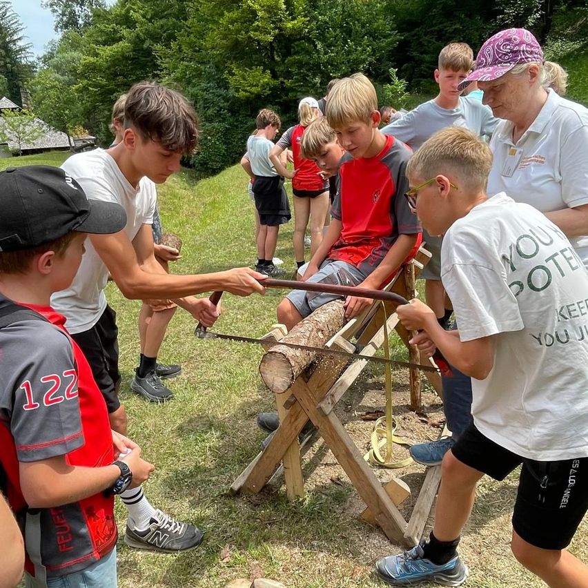 Eine Gruppe von Kindern und Erwachsenen ist im Freien und säg einen Baumstamm. Die Kinder tragen Turnschuhe und einige tragen Mützen. Die Erwachsenen beobachten sie.