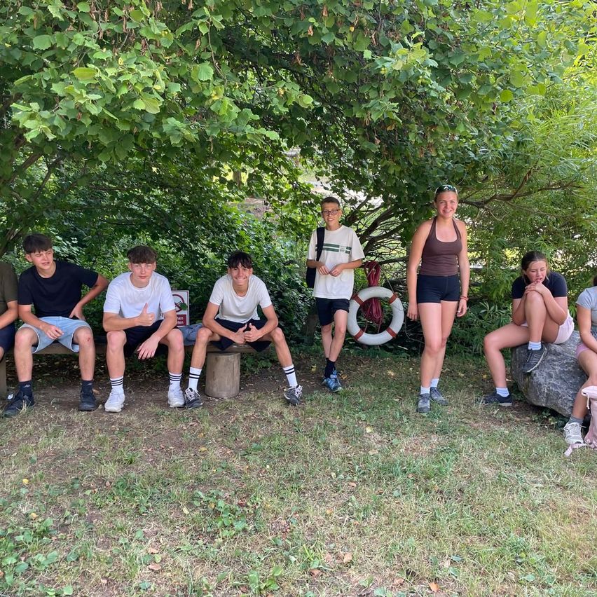 Eine Gruppe junger Leute posiert für ein Foto unter einem Baum in einem Park, einige sitzen auf einer Bank und andere stehen. Ein Rettungsring hängt am Baum.