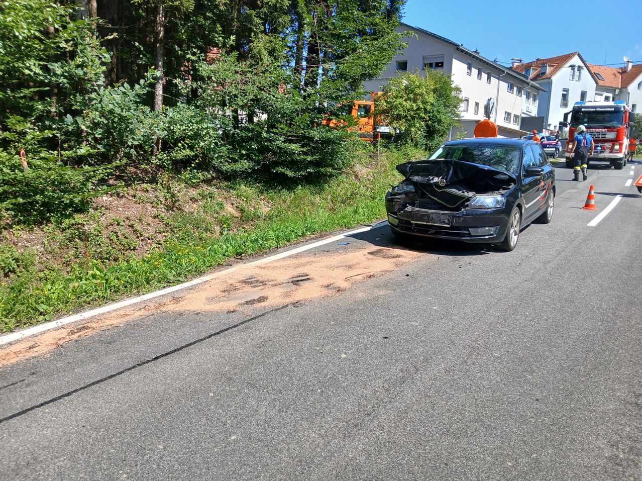 Ein schwarzer Wagen mit geöffneter Motorhaube steht am Straßenrand, mit einem Verkehrskegel darauf. Die Straße ist von Grünflächen und einem Gebäude umgeben.