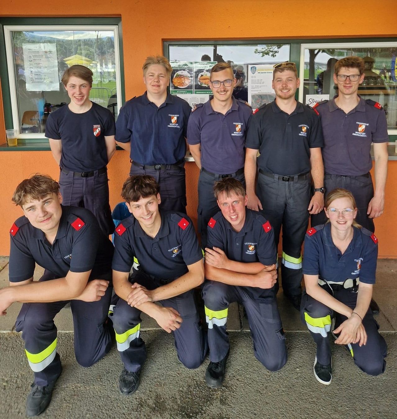 Eine Gruppe von Feuerwehrleuten in Uniform, die vor einem Gebäude mit Fenstern und einer Glastür für ein Foto posieren.