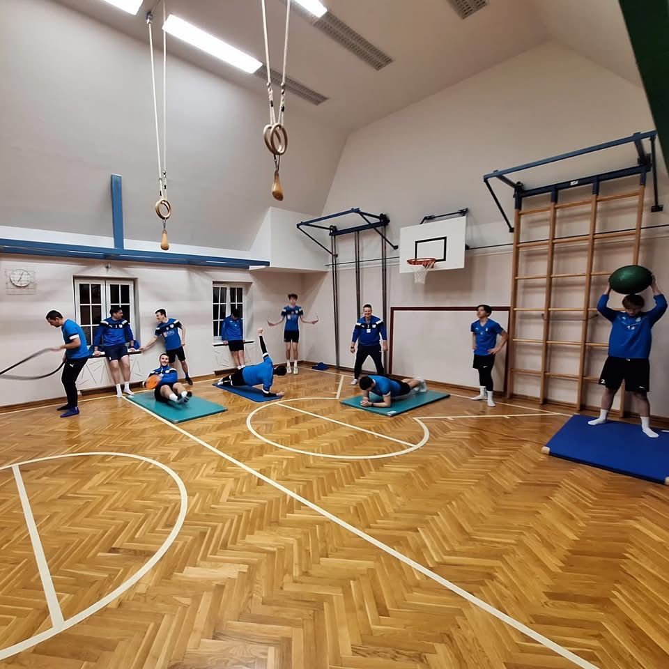 Eine Gruppe von Menschen in blauen Uniformen trainiert in einer Turnhalle mit einem Basketballplatz. Einige sind auf dem Boden mit Trainingsmatten. Geräte und ein Basketballkorb sind sichtbar.