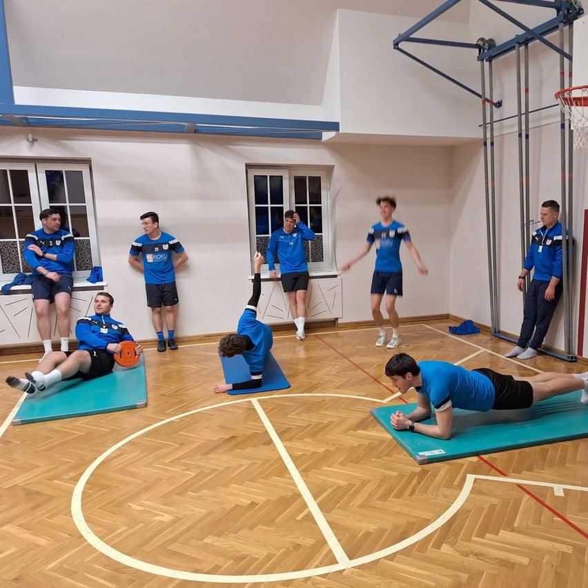 Eine Gruppe von Männern in blauen Uniformen macht körperliche Übungen in einer Turnhalle. Einige sitzen auf Matten, während andere stehen. Ein Basketballkorb befindet sich auf der rechten Seite.
