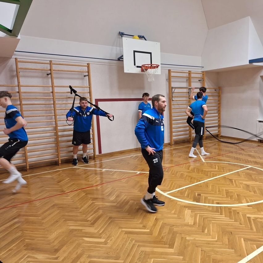 Mehrere Männer machen Übungen mit Widerstandsbändern auf einem Holzboden einer Indoor-Basketballhalle. Ein Mann läuft, während ein anderer anweist.