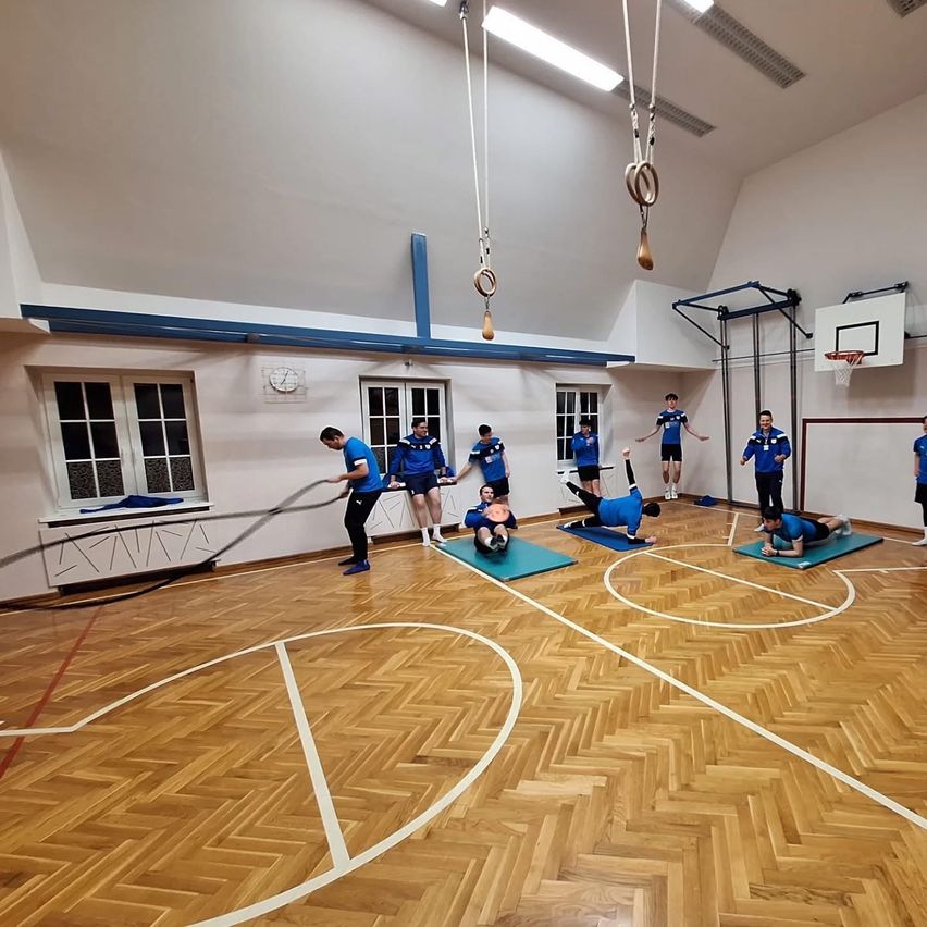 Innengymnasium mit Holzboden, eine Gruppe von Personen macht Übungen mit Matten, zwei machen Klimmzüge an Ringen, einer benutzt eine schwere Schnur.