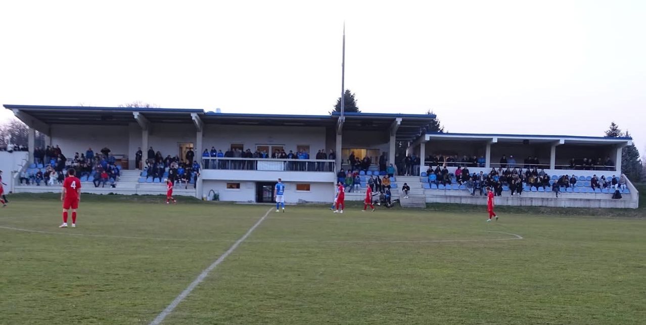Ein Fußballspiel findet auf einem Spielfeld statt, das von einer großen Menge aus den Tribünen beobachtet wird. Die Spieler tragen rote und blaue Uniformen.