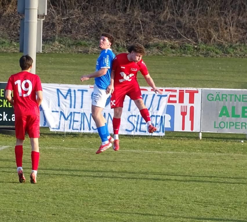 Zwei Fußballspieler in Aktion, einer trägt ein blaues Trikot und der andere ein rotes, sind in der Luft, möglicherweise in einem Wettkampf. Ein dritter Spieler in Rot steht hinter ihnen auf der linken Seite.