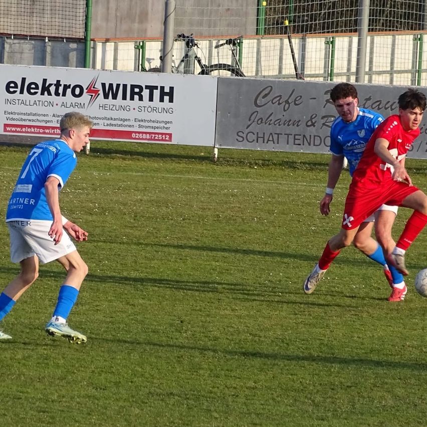 Drei Fußballspieler sind auf einem Feld. Einer in Blau jagt den Ball, während zwei in Rot verteidigen. Dahinter ist eine Werbung für Elektro Wirth zu sehen.