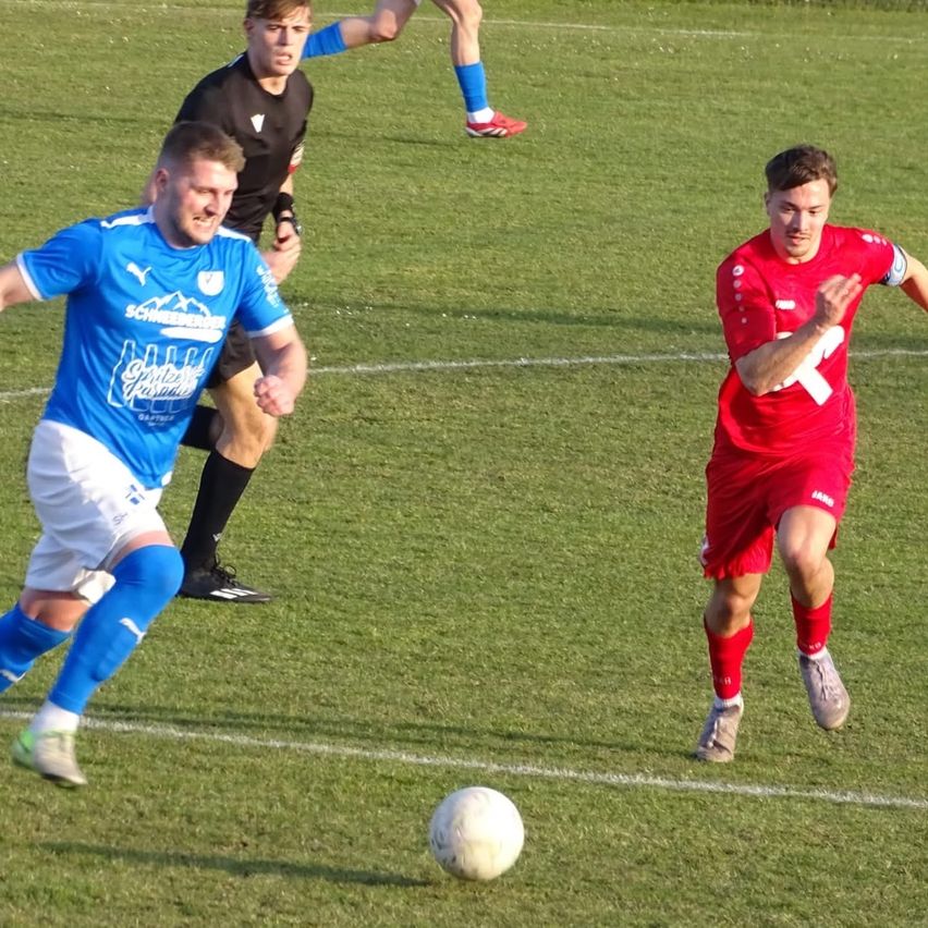Drei Fußballspieler auf einem Feld, einer in Blau, einer in Rot und einer in Schwarz, jagen einem Ball hinterher.