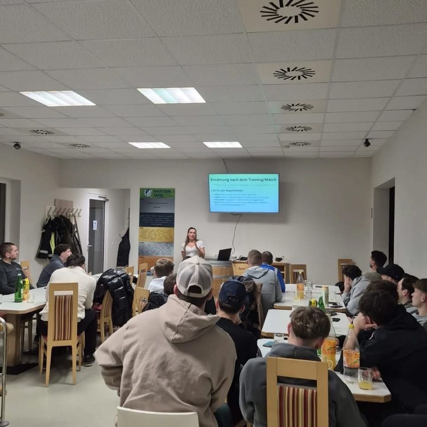 Eine Frau steht vor einer Projektionsleinwand in einem Klassenzimmer. Mehrere Personen sitzen an Tischen vor ihr und lauschen aufmerksam.