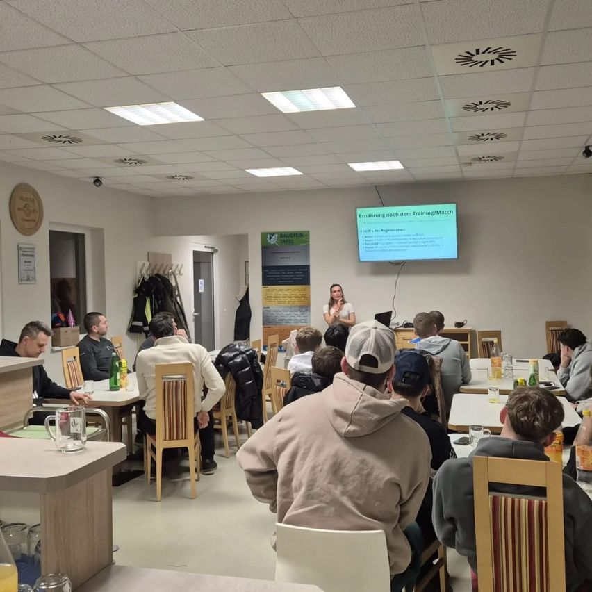 Eine Frau hält eine Präsentation vor einem Raum. Mehrere Personen sitzen um Tische herum und lauschen. Eine Projektionsleinwand ist hinter der Frau sichtbar.