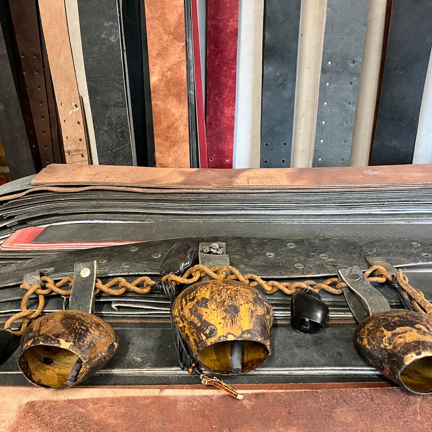 Verschiedene Ledermaterialien sind im Hintergrund zu sehen. Einige Ledergürtel sind übereinander gestapelt. Ein paar Glocken sind mit einer Kette vorne verbunden.