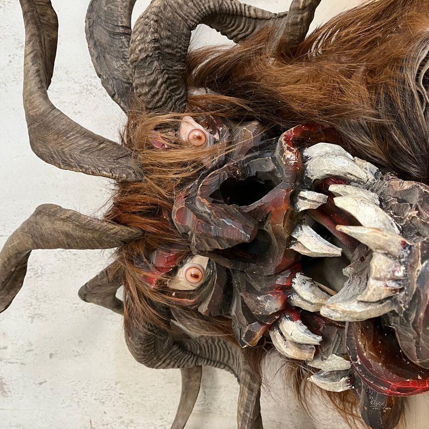 Nahaufnahme einer großen gehörnten und haarigen Maske mit weit geöffnetem Mund, vor weißem Hintergrund.