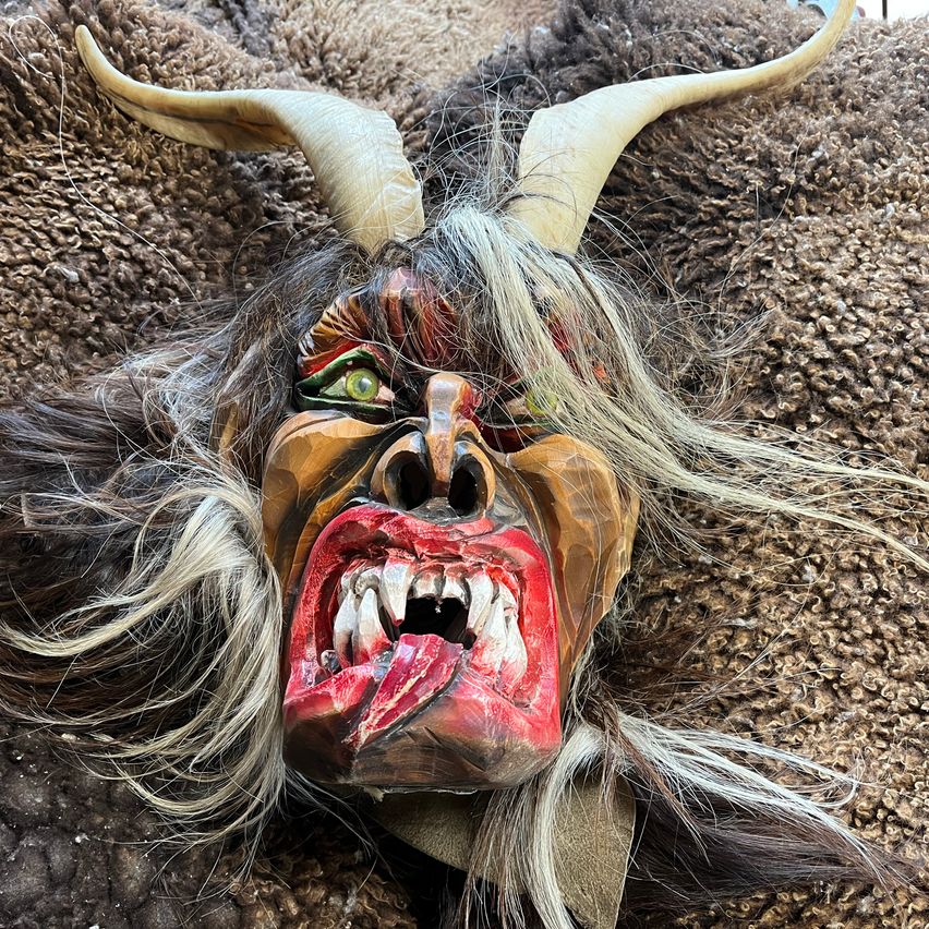 Eine hölzerne Maske mit Hörnern und wildem Haar liegt auf einer braunen Oberfläche. Die Maske hat einen großen offenen Mund mit Zähnen und grünen Augen.