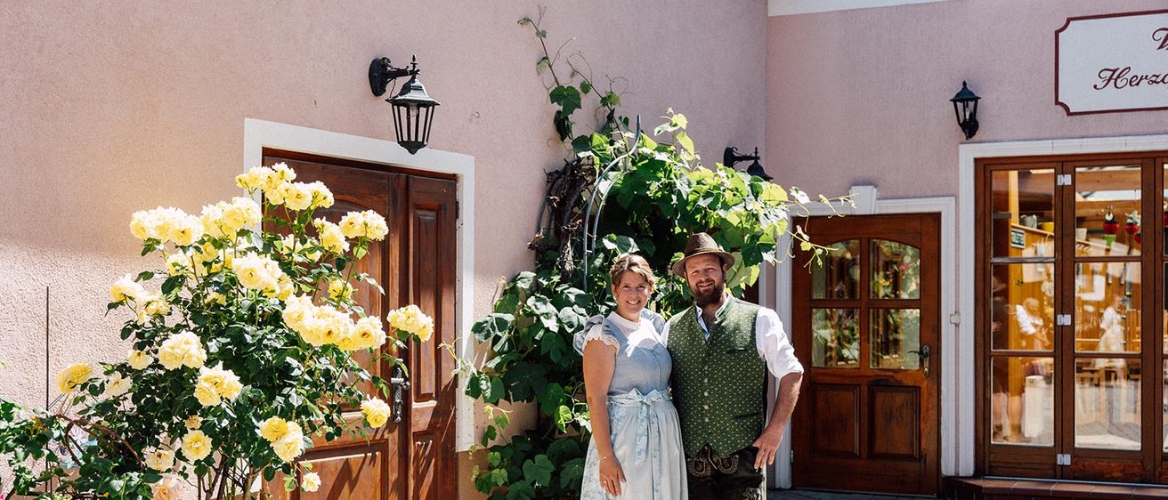 Ein Mann und eine Frau in traditioneller Kleidung stehen vor einem Haus mit zwei Holztüren. Das Haus hat eine rosa Wand und es gibt gelbe Blumen, eine Pflanze und eine Lampe an der Wand.