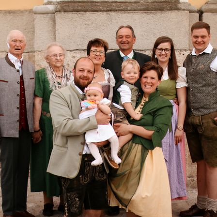 Eine Familie in traditioneller Kleidung posiert für ein Foto mit zwei kleinen Kindern. Die Umgebung ist draußen, und alle lächeln.