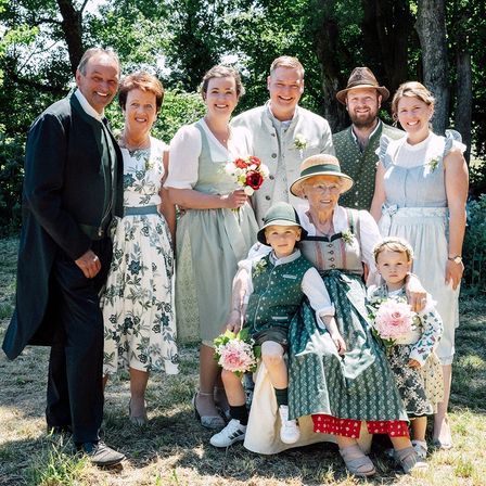 Eine Familie von sieben Personen in traditioneller deutscher Kleidung posiert für ein Foto im Garten. Eine ältere Frau hält Blumen, ein Kind sitzt auf ihrem Schoß und andere stehen hinter ihnen.