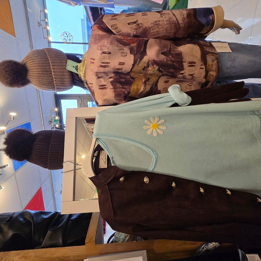 Eine Puppe trägt eine braune Jacke und Mütze, darunter ein hellblaues Sweatshirt und eine braune Wildlederjacke. Die Szene umfasst eine weiße Vitrine und eine Wand mit blau-roter Musterung.