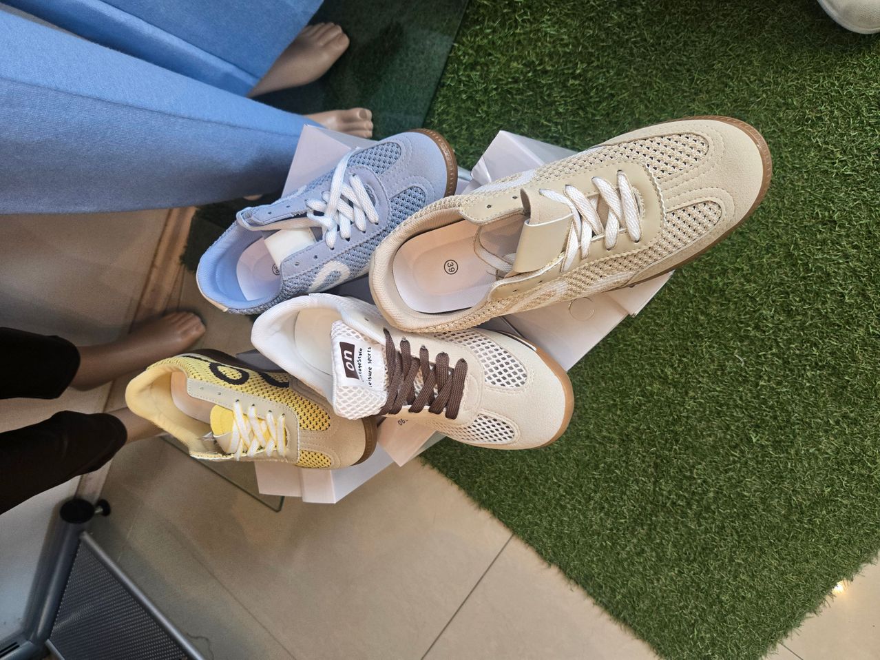 A display of four sneakers in various colors, including blue, beige, white, and yellow, rests on a green carpet. The sneakers are placed on white boxes.