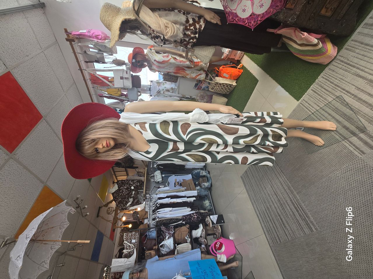 A mannequin wearing a red hat and a patterned dress is displayed in a clothing store with various items on shelves and a green carpet on the floor.