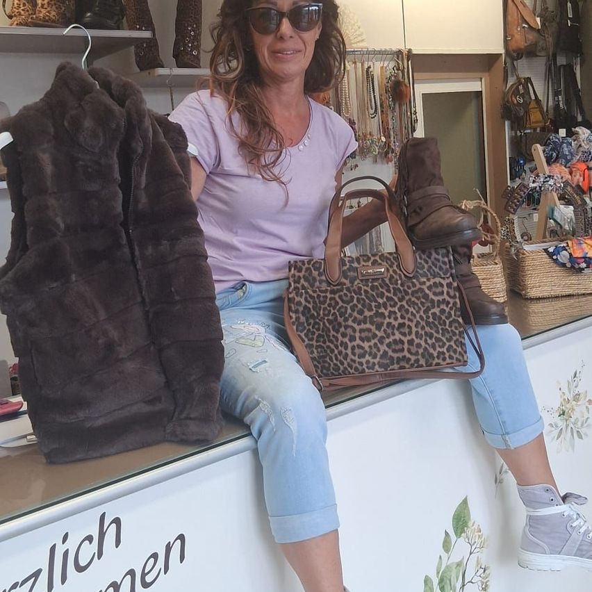 A woman sits on a counter in a clothing store, holding a brown fur vest and a leopard print handbag. She wears sunglasses and sneakers. Behind her, shelves display various items, including bags and jewelry.