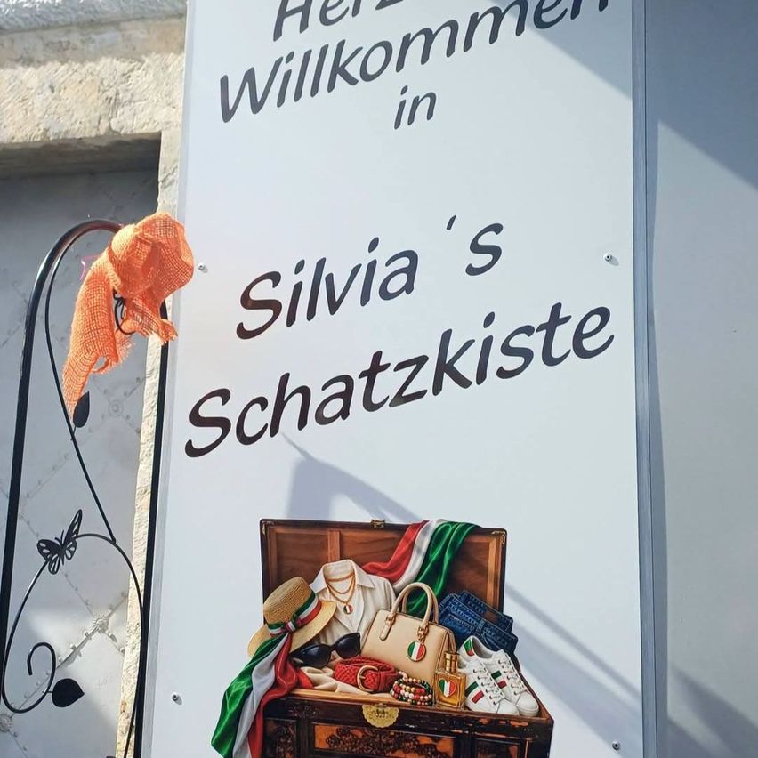 A sign with a trunk full of fashion items, including hats, bags, and shoes, reads 'Herzlich Willkommen in Silvia's Schatzkiste'.
