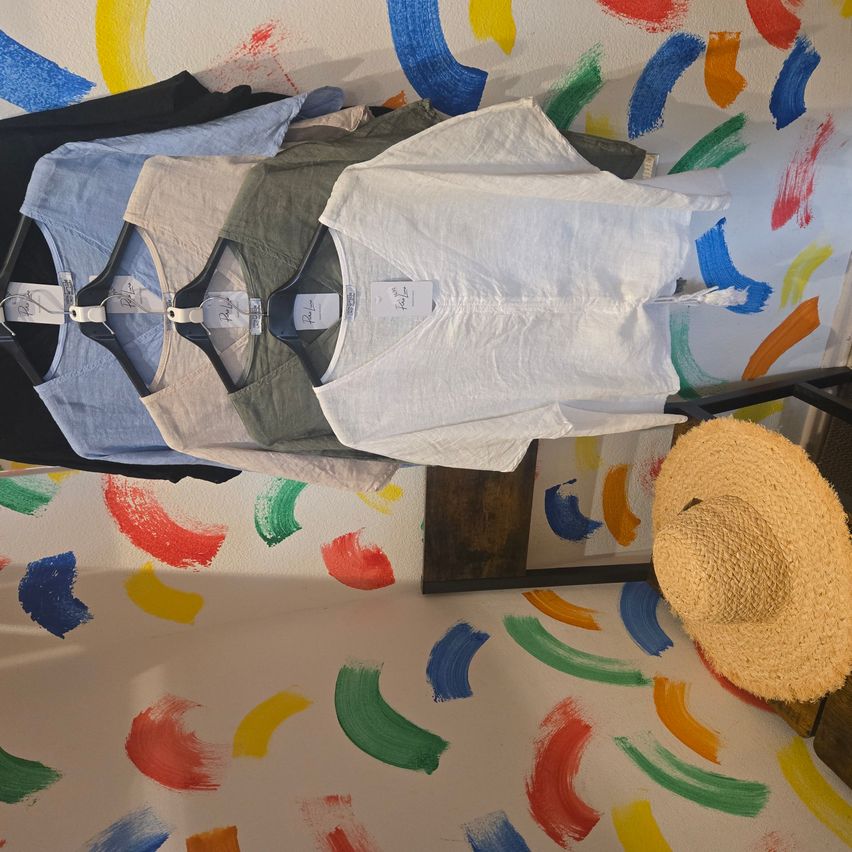 Verschiedene T-Shirts hängen an einem Ständer an einer Wand mit bunten Farbspritzern. Ein Strohhut liegt neben dem Ständer auf dem Boden.