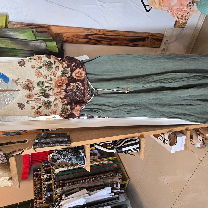 Ein floral bedrucktes Oberteil und grüne Hosen an einem Ständer in einem Bekleidungsgeschäft mit verschiedenen Artikeln wie Handtaschen und Schuhen.
