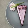 Ein Stück Kuchen mit rosa Zuckerguss und Kräutern auf einem grauen Teller, begleitet von einer rosa Blume mit grünen Blättern.