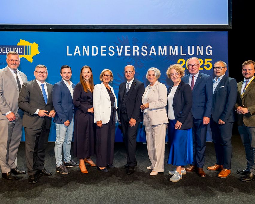 Eine Gruppe von Menschen posiert für ein Foto vor einem blauen Hintergrund mit 'Landesversammlung 2025' darauf. Sie sind alle formell gekleidet.