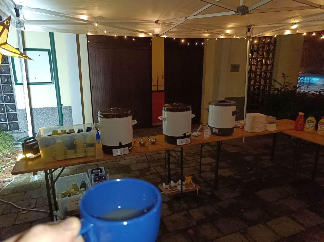 Ein Innenbereich mit einem Tisch, auf dem drei elektrische Wasserkocher und eine blaue Tasse stehen. Der Raum hat Holzböden, eine Zeltdecke mit Lichtern und große Holztüren.