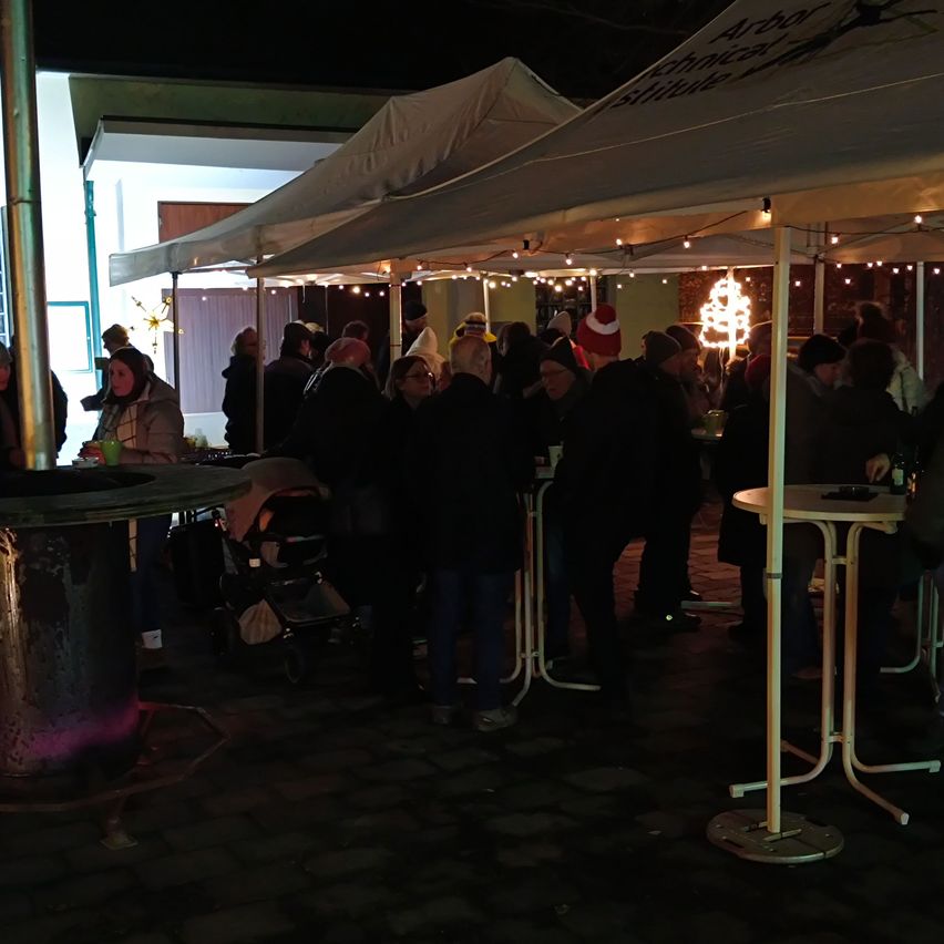 Bei Nacht versammeln sich Menschen unter Zelten mit Lichterketten. Einige tragen Mützen und Brillen. Tische sind mit Flaschen und Bechern bestückt. Ein Kinderwagen steht in der Nähe.