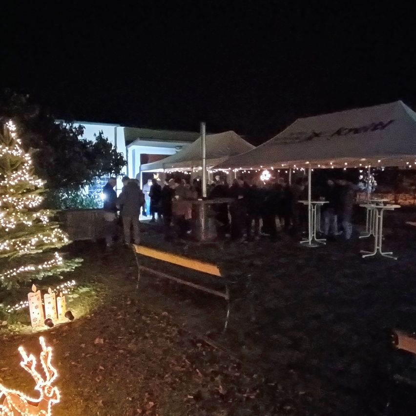 Eine Gruppe von Menschen versammelt sich unter zwei Zelten mit Tischen und Stühlen, beleuchtet von Lichterketten. Ein mit Lichtern geschmückter Weihnachtsbaum steht links.