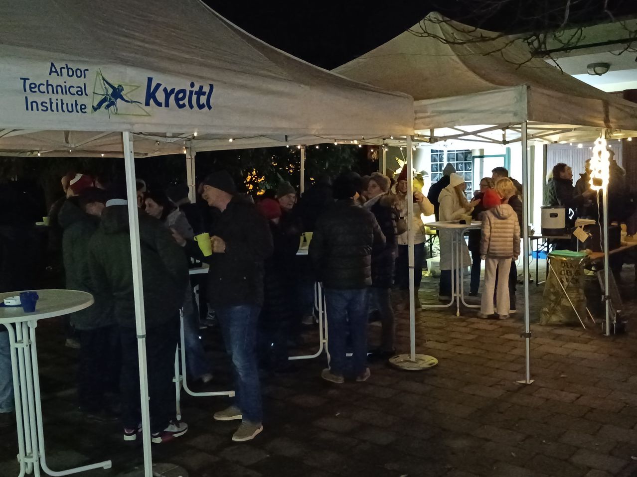 Menschen stehen unter weißen Zelten bei einer nächtlichen Veranstaltung Schlange und halten Tassen. Die Zelte sind mit 'Kreitl' beschriftet.