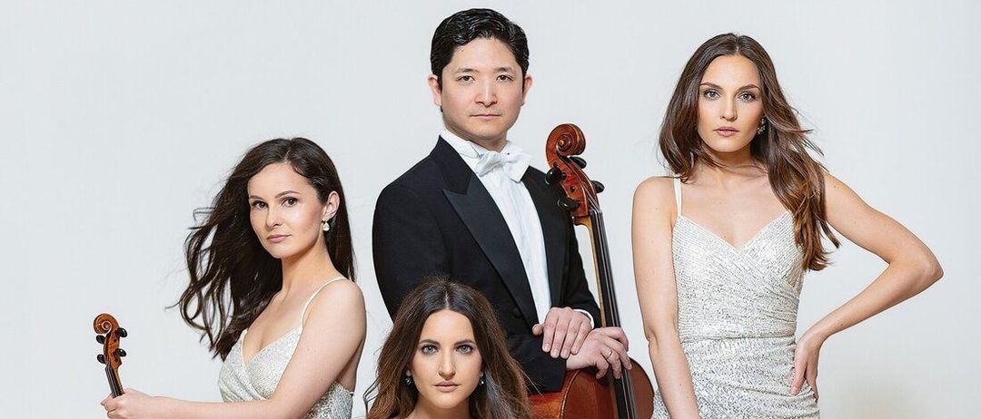 Eine Gruppe von Musikern, drei Frauen und ein Mann, posiert für ein Foto. Sie sind in formeller Kleidung gekleidet und halten Saiteninstrumente. Der Mann hält eine Geige, während die Frauen Violinen halten. Sie alle schauen in die Kamera.