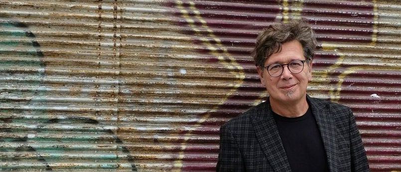 Ein Mann mit einer Brille und einem karierten Anzug steht vor einer mit Graffiti bedeckten Wand. Er lächelt.