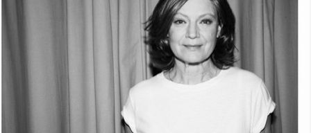 Ein Schwarz-Weiß-Porträt einer Frau mit kurzen Haaren, sie lächelt und trägt ein weißes T-Shirt. Sie steht vor einem unscharfen Hintergrund aus Vorhängen.