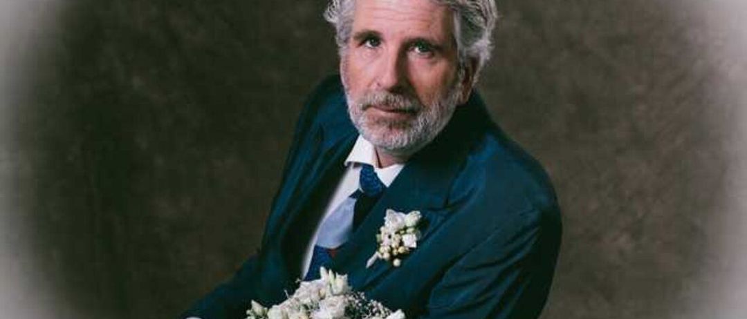 Ein älterer Mann mit grauem Haar und Bart trägt einen dunklen Anzug, eine Krawatte und hält einen weißen Blumenstrauß.