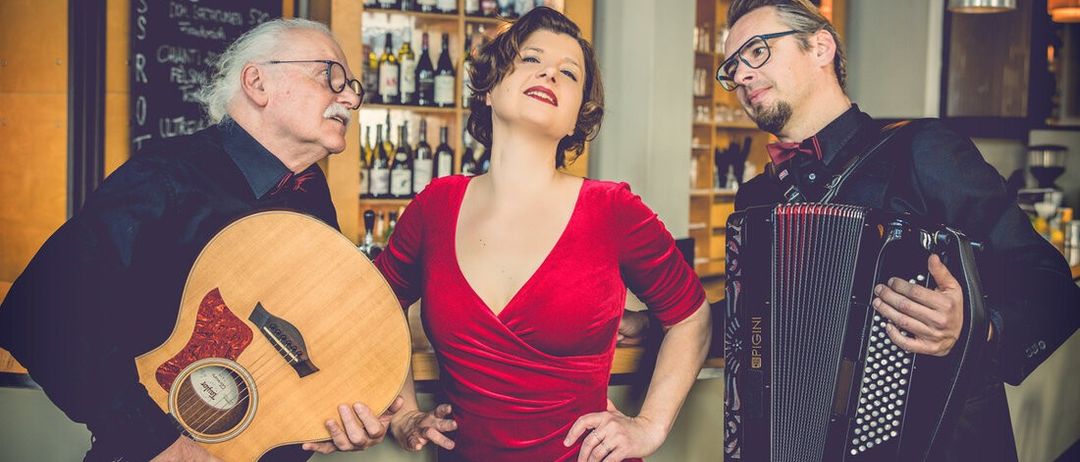 Eine Frau in einem roten Kleid steht zwischen zwei Männern, einer hält eine Gitarre und der andere spielt Akkordeon. Sie alle lächeln und scheinen für ein Foto in einer Bar zu posieren.