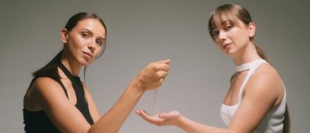 Zwei Frauen sitzen an einem Tisch. Eine hält eine Sanduhr. Der Text darüber lautet 'Piano & Sand'. Die Frauen werden als Anna Vidyakina und Sabina Hasanova identifiziert.