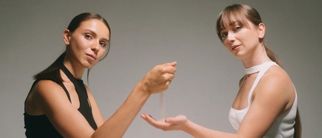 Zwei Frauen sitzen an einem Tisch. Eine hält eine Sanduhr. Der Text darüber lautet 'Piano & Sand'. Die Frauen werden als Anna Vidyakina und Sabina Hasanova identifiziert.